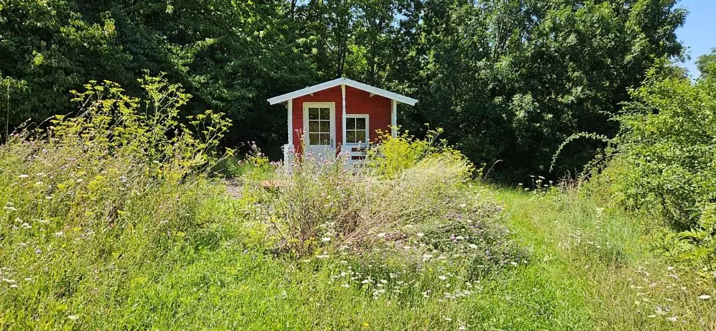 Kleines rotes Holzhaus im Seminargarten von Maria Richter, umgeben von hoher Blumenwiese und Bäumen.